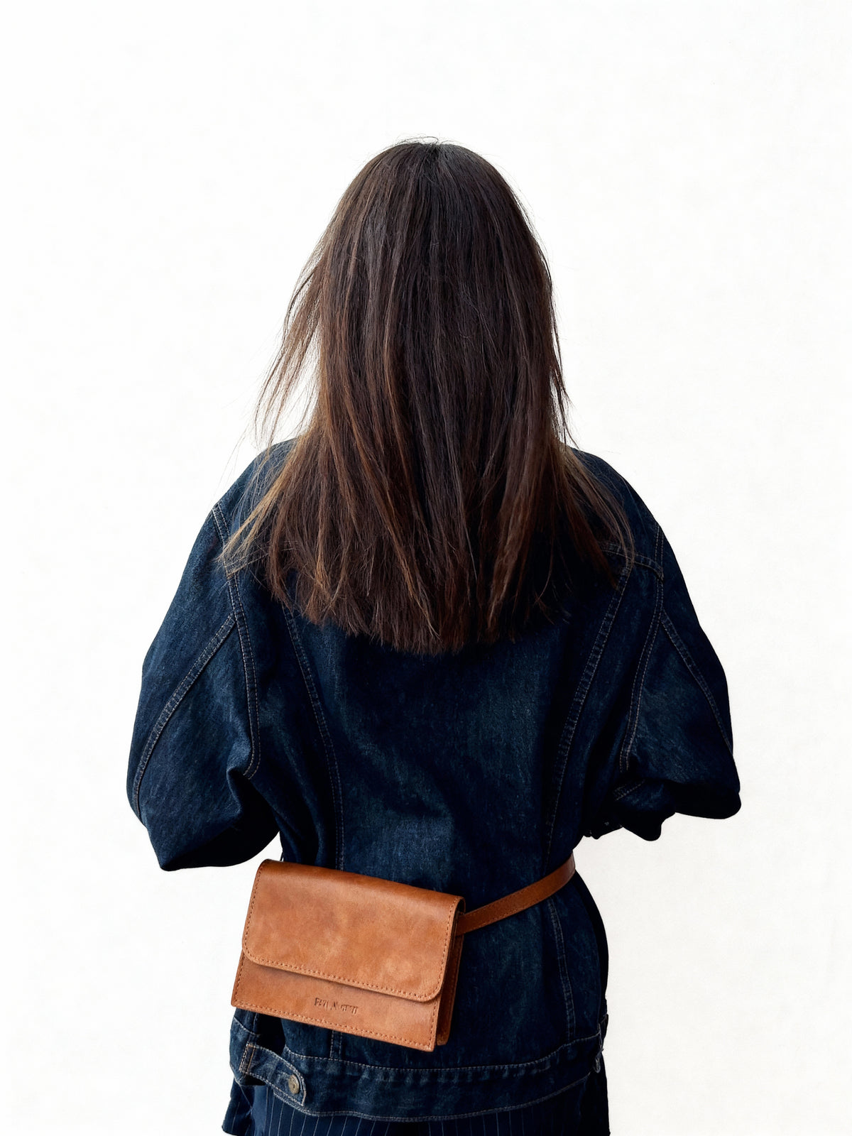 Person wearing a denim jacket with a brown leather waist bag on a white background