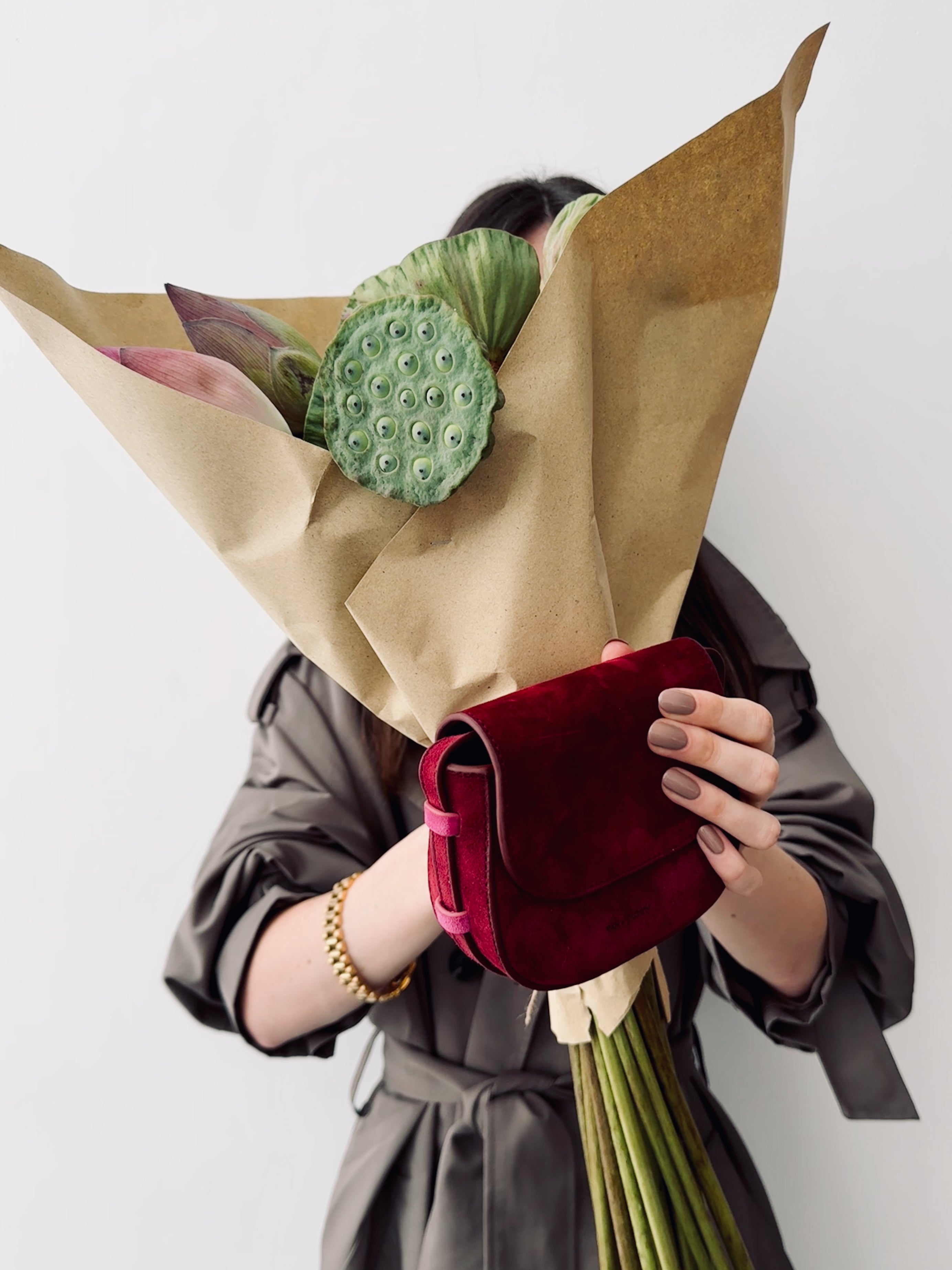 Person holding a bouquet of flowers wrapped in brown paper with a red bag underneath, on a white background.