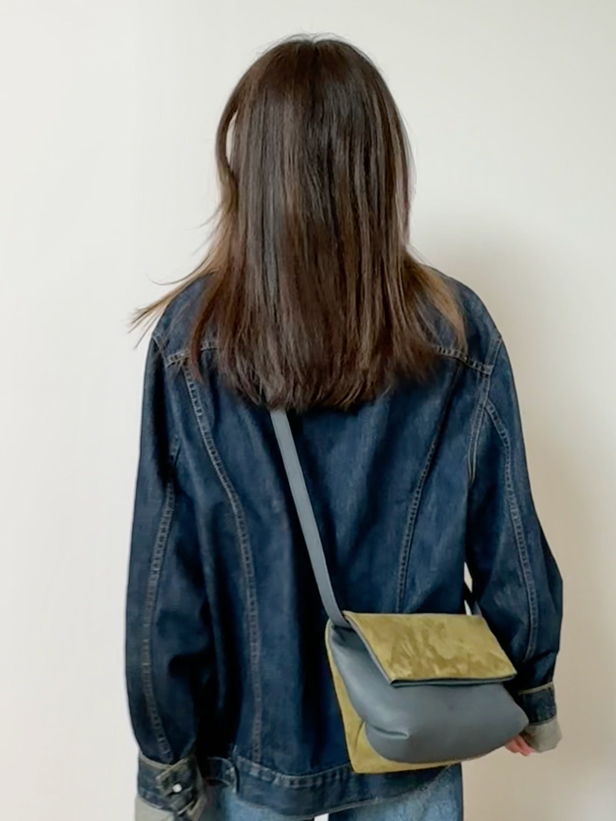Person wearing a denim jacket with a green and gray crossbody bag on a plain background